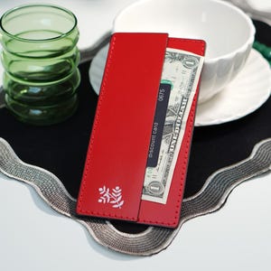 May include: A rectangular red leather card holder featuring a white botanical sprig icon on the front. The card holder contains a black discount card and a one-dollar bill tucked into the slot. The edges are stitched with matching red thread.