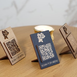 Könnte beinhalten: Drei Menükartenhalter aus Holz mit Rosenmuster und QR-Codes. Die Halter sind in Hellbraun, Dunkelblau und Dunkelbraun gehalten. Jeder Halter trägt unten den Text "SCAN FOR MENU". Sie werden auf einem Holztisch präsentiert.
