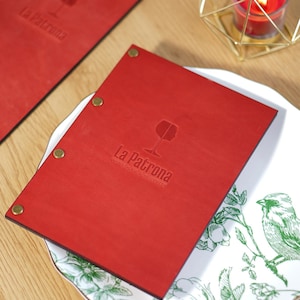 May include: Two rectangular red menu covers with brass-colored metal fasteners. Each cover features an embossed wine glass icon and the text La Patrona Wine Bar & Bistro. The covers are placed on a white plate with a green botanical bird pattern.