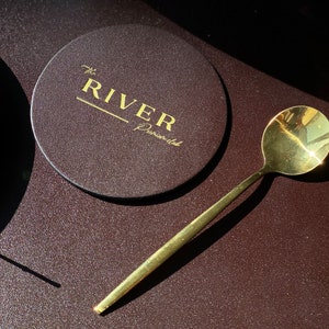 Può includere: Un sottobicchiere in pelle marrone con lettere dorate che recitano "The River, Peninsula". Un cucchiaio d'oro giace su una superficie marrone testurizzata.