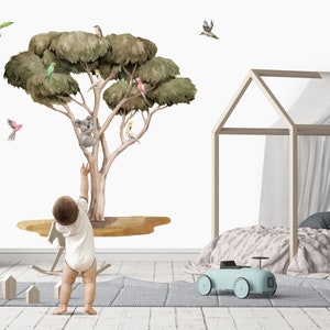 Op de afbeelding: Een muursticker in aquarelstijl met een eucalyptusboom met een koala, vogels en een gele vogel. De sticker is te zien op een witte muur in een kinderkamer met een kind er voor.