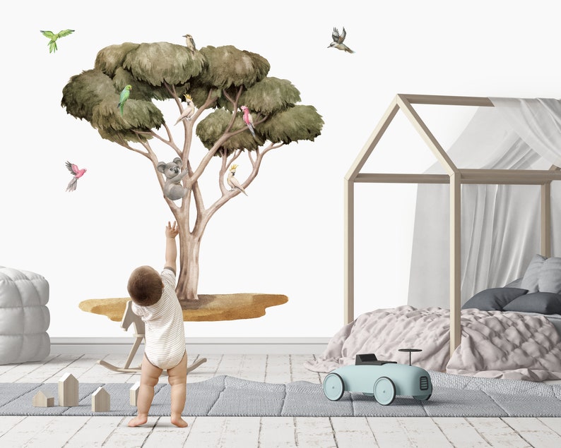 Op de afbeelding: Een muursticker in aquarelstijl met een eucalyptusboom met een koala, vogels en een gele vogel. De sticker is te zien op een witte muur in een kinderkamer met een kind er voor.