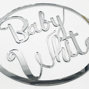 Könnte beinhalten: Ein silberner Spiegelkreis mit den Worten "Baby White" in kursivschrift.