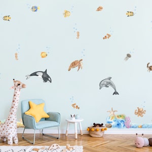 Peut inclure: Un mur bleu décoré d'une variété d'autocollants d'animaux marins colorés, dont un dauphin, une tortue, un crabe, une méduse, un hippocampe et un poisson. On y voit également un animal en peluche en forme de girafe et un fauteuil bleu avec un coussin jaune.