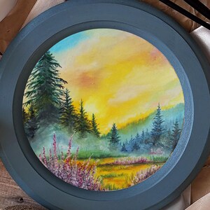 Puede incluir: Pintura de paisaje enmarcada, redonda, que representa una escena de bosque. La obra presenta una puesta de sol con tonos amarillos y anaranjados, árboles verdes y flores silvestres moradas. Una vela encendida, pinceles y una cinta están cerca.