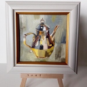 Puede incluir: Una pequeña pintura al óleo de una tetera plateada con tapa y asa doradas, sobre un fondo tenue. La pintura está enmarcada en un marco blanco con un borde interior dorado. La pintura está sobre un pequeño caballete de madera con la palabra "ART" impresa en él.