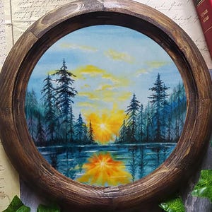 Puede incluir: Pintura redonda en acuarela enmarcada en madera que representa una puesta de sol sobre un lago. La obra presenta un sol amarillo y naranja brillante que se pone detrás de árboles oscuros, reflejado en el agua. El marco es de color marrón oscuro.