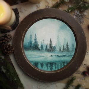 Puede incluir: Una pintura redonda enmarcada en un marco marrón oscuro muestra una escena de bosque brumoso con árboles de hoja perenne reflejados en el agua. El fondo incluye velas, piñas y partituras.