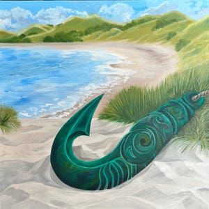 May include: A green, carved, wooden hook, known as a 'hei tiki', lies on a sandy beach with green grass and a blue ocean in the background.
