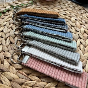 May include: A collection of corduroy keychains in various colors, including brown, blue, green, gray, and pink. Each keychain has a metal ring and a fabric loop. The keychains are arranged on a woven, textured surface.
