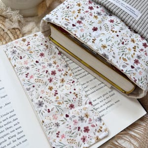 Eine Stoffbuchhülle mit weißem Hintergrund und einem zarten Blumenmuster in Rosa-, Rot- und Grüntönen. Die Hülle hat einen gerippten grauen oberen Teil mit einem weißen Etikett inklusive Lesezeichen.