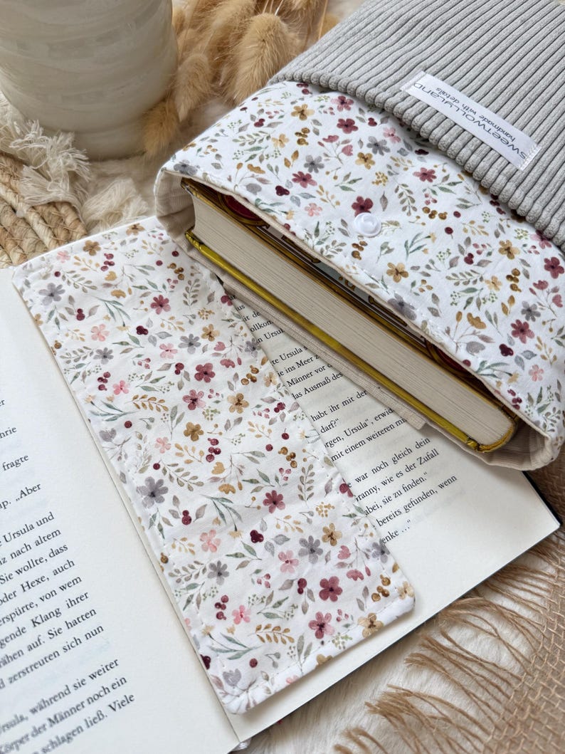 Eine Stoffbuchhülle mit weißem Hintergrund und einem zarten Blumenmuster in Rosa-, Rot- und Grüntönen. Die Hülle hat einen gerippten grauen oberen Teil mit einem weißen Etikett inklusive Lesezeichen.