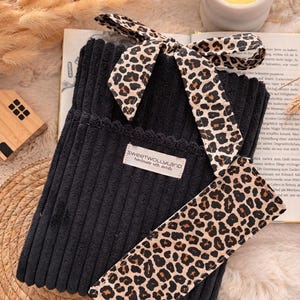 Könnte beinhalten: Schwarze Cord-Tasche mit Leopardenmuster-Schleife und passendem Riemen. Die Tasche hat einen gewellten Rand und ein Etikett mit der Aufschrift "SWEETWOLLYLAND handmade with details". Eine brennende Kerze und ein Buch sind im Hintergrund.