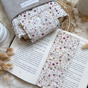 Könnte beinhalten: Eine graue Buchhülle aus cord mit einem passenden Lesezeichen mit Blumenmuster die Buchschutz  liegt auf einem aufgeschlagenen Buch mit deutschem Text.