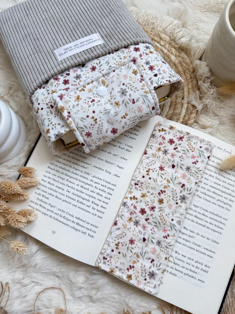 Könnte beinhalten: Eine graue Buchhülle aus cord mit einem passenden Lesezeichen mit Blumenmuster die Buchschutz  liegt auf einem aufgeschlagenen Buch mit deutschem Text.