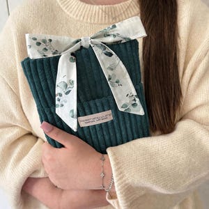 Könnte beinhalten: Eine tealgrüne Cord-Tasche mit einem weißen Band mit grünem Blättermuster. Die Tasche wird von einer Person gehalten, die einen cremefarbenen Pullover trägt. Der Text "sweetwolluland handmade with details" steht auf einem kleinen Etikett.