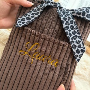 Peut inclure: Une housse de bouillotte en velours c&ocirc;tel&eacute; marron avec une surface textur&eacute;e &agrave; c&ocirc;tes et un n&oelig;ud d&eacute;coratif en tissu &agrave; motif l&eacute;opard nou&eacute; sur le dessus. Le pr&eacute;nom Laura est brod&eacute; en fil dor&eacute; cursif sur le devant de la housse en tissu marron.