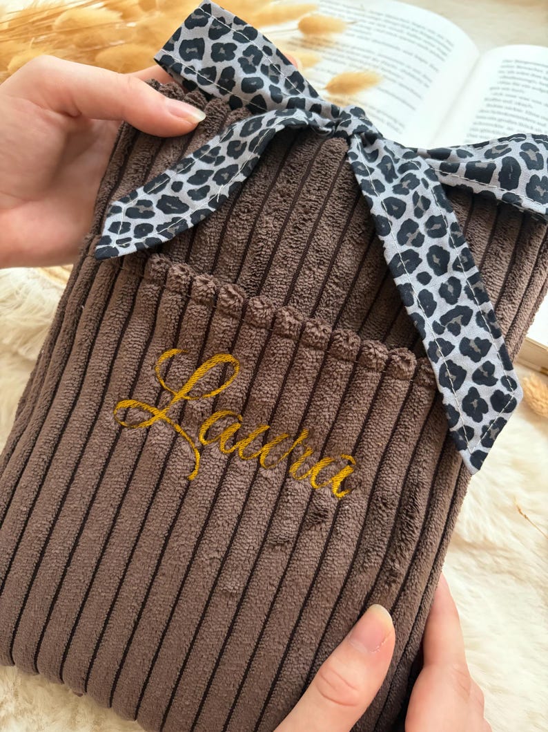 Peut inclure: Une housse de bouillotte en velours c&ocirc;tel&eacute; marron avec une surface textur&eacute;e &agrave; c&ocirc;tes et un n&oelig;ud d&eacute;coratif en tissu &agrave; motif l&eacute;opard nou&eacute; sur le dessus. Le pr&eacute;nom Laura est brod&eacute; en fil dor&eacute; cursif sur le devant de la housse en tissu marron.