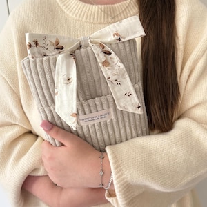 Könnte beinhalten: Ein beiger Cord-Buchumschlag mit einer floral gemusterten Schleife. Die Schleife besteht aus cremefarbenem Stoff mit braunen und beigen floralen Designs. Der Buchumschlag hat ein Etikett mit der Aufschrift "sweetwollyland handmade with details".