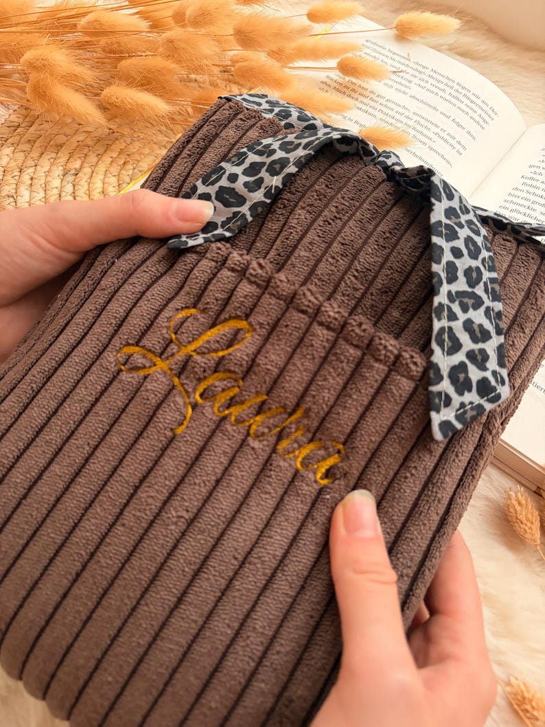 Peut inclure: Une housse de bouillotte en velours c&ocirc;tel&eacute; marron avec une surface textur&eacute;e &agrave; c&ocirc;tes et un lien en tissu &agrave; motif l&eacute;opard sur le dessus. Le pr&eacute;nom "Laura" est brod&eacute; en fil dor&eacute; cursif sur le devant de la housse, qui comporte une petite poche.