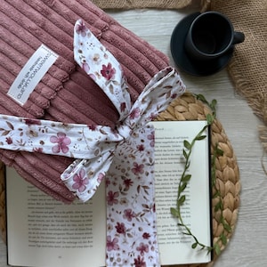 Könnte beinhalten: Eine staubrosa Cord-Buchhülle mit floralem Schleifenband und passendem Lesezeichen. Lesezeichen und Band zeigen einen weißen Hintergrund mit bordeauxroten und rosa Blumenmustern. Das Buch ist auf einer gewebten Matte geöffnet.