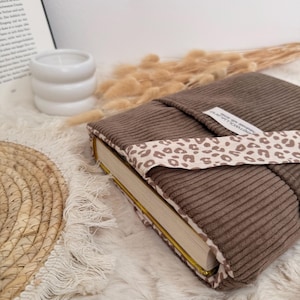 Ein brauner Cord-Buchumschlag mit einem Leopardenmusterband. Der Buchumschlag ist um ein Buch mit goldfarbenen Seiten gewickelt. Der Hintergrund umfasst eine weiße Kerze, Trockenblumen und eine gewebte Matte.