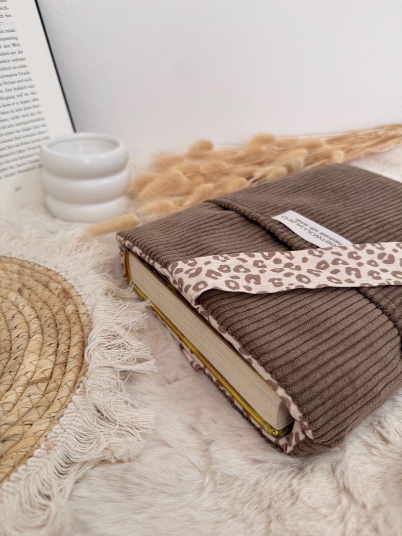 Ein brauner Cord-Buchumschlag mit einem Leopardenmusterband. Der Buchumschlag ist um ein Buch mit goldfarbenen Seiten gewickelt. Der Hintergrund umfasst eine weiße Kerze, Trockenblumen und eine gewebte Matte.