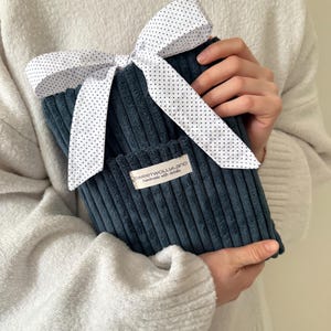 Könnte beinhalten: Eine tealfarbene Cord-Wärmflaschenhülle mit einer weißen Schleife mit Polka-Dots. Die Hülle hat eine kleine Tasche und ein Etikett mit der Aufschrift "sweetwollyland handmade with details". Der Artikel wird von einer Person gehalten, die einen weißen Pullover trägt.