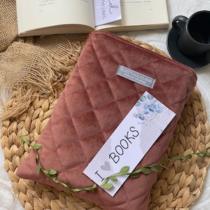 Könnte beinhalten: Ein gesteppter, staubrosafarbener Buchumschlag mit einem "SWEETWOLLYLAND"-Etikett und einem Lesezeichen mit der Aufschrift "Read BETWEEN THE LINES". Ein Lesezeichen mit "I BOOKS" ist ebenfalls vorhanden. Der Buchumschlag liegt auf einer gewebten Matte.