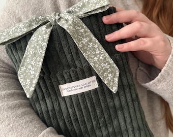 Peut inclure: Une pochette en velours côtelé vert foncé, ornée d'un nœud à motifs floraux blancs et verts. L'étiquette indique "sweetwollyland handmade with details". La pochette est tenue par une personne portant un pull beige. Le tissu est côtelé.