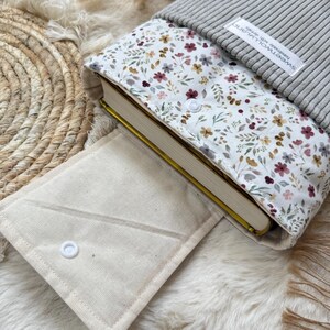 Eine Buchhülle aus Stoff mit weißem Blumenmuster aus kleinen roten, gelben und grünen Blüten, einem grauen, gerippten Strickoberteil und einer beigen Canvas-Klappe mit weißem Druckknopf. Ein weißes Etikett trägt die Aufschrift sweetwolluland handmade with details.