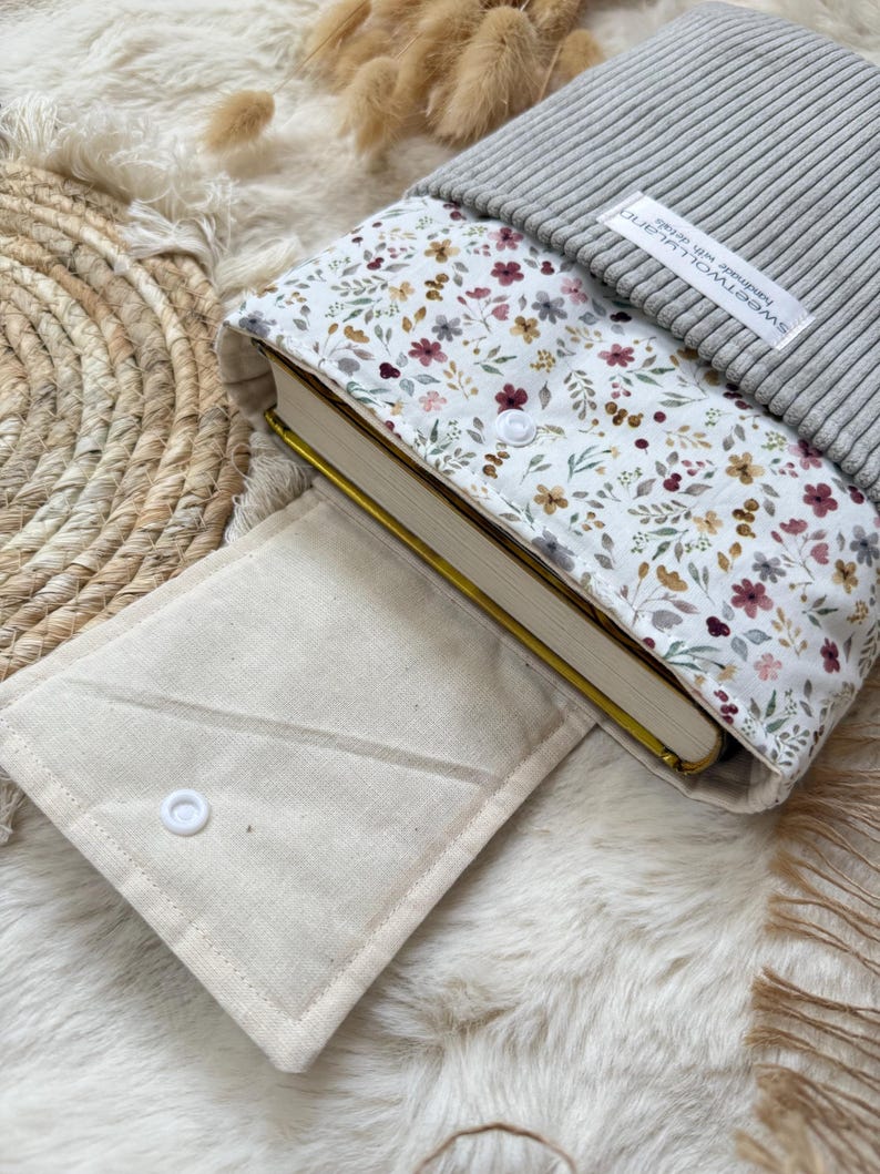 Eine Buchhülle aus Stoff mit weißem Blumenmuster aus kleinen roten, gelben und grünen Blüten, einem grauen, gerippten Strickoberteil und einer beigen Canvas-Klappe mit weißem Druckknopf. Ein weißes Etikett trägt die Aufschrift sweetwolluland handmade with details.