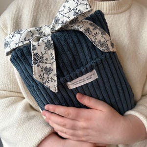 Könnte beinhalten: Eine marineblaue Cord-Wärmflasche, gebunden mit einem cremefarbenen Band mit schwarzen Blumenmustern. Das Etikett lautet "SWEETWOLLYLAND handmade with details."