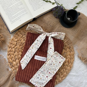 Könnte beinhalten: Ein Buchhülle aus braunem Cord, kombiniert mit einem passenden Stirnband und Armband mit floralem Muster. Stirnband und Armband haben einen zarten Blumendruck auf weißem Hintergrund. Auf der Buchhülle steht "sweetwoolland handmade with details."
