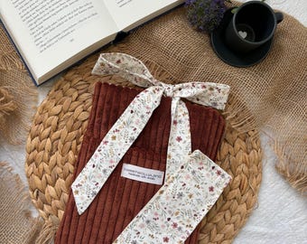 May include: A corduroy book sleeve in a rich brown hue, paired with a matching floral patterned headband and wristband. The headband and wristband feature a delicate floral print on a white background. The book sleeve has a tag that reads "sweetwoolland handmade with details."