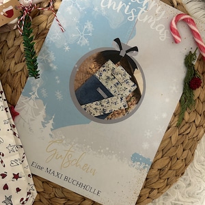 May include: A Christmas-themed gift certificate with a blue and white background, featuring a decorative ornament design. The ornament contains fabric book covers. The words "merry christmas" are at the top. The text "Gutschein" and "Eine MAXI BUCHHÜLLE" are also visible.