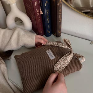 Ein brauner Buchumschlag mit Cordstruktur und einem Schleifenband mit Leopardenmuster. Das Kissen hat ein kleines weißes Etikett mit Text. Dekorative Bücher mit goldener Schrift und eine weiße Vase befinden sich im Hintergrund.