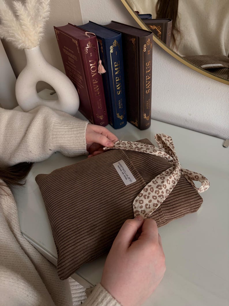 Ein brauner Buchumschlag mit Cordstruktur und einem Schleifenband mit Leopardenmuster. Das Kissen hat ein kleines weißes Etikett mit Text. Dekorative Bücher mit goldener Schrift und eine weiße Vase befinden sich im Hintergrund.