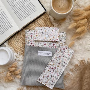 Ein Buch mit einem floralen Lesezeichen, einer grauen Cord-Buchhülle und einer weißen Tasse. Das Lesezeichen und die Oberseite der Buchhülle haben einen weißen Hintergrund mit einem floralen Muster in Rosa-, Braun- und Grüntönen. Die Buchhülle trägt den Text "sweetwollyland handmade with details".