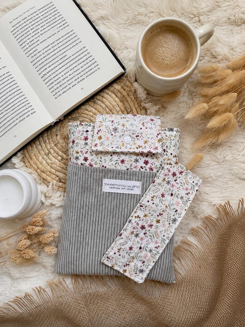 Ein Buch mit einem floralen Lesezeichen, einer grauen Cord-Buchhülle und einer weißen Tasse. Das Lesezeichen und die Oberseite der Buchhülle haben einen weißen Hintergrund mit einem floralen Muster in Rosa-, Braun- und Grüntönen. Die Buchhülle trägt den Text "sweetwollyland handmade with details".