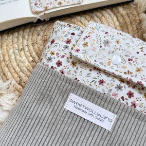 Ein grauer Beutel aus Rippenstoff mit einer blumig gemusterten Klappe und einem weißen Druckknopfverschluss. Auf der Vorderseite ist ein weißes rechteckiges Etikett mit dem schwarzen Text "sweetwollyland handmade with details" aufgenäht.