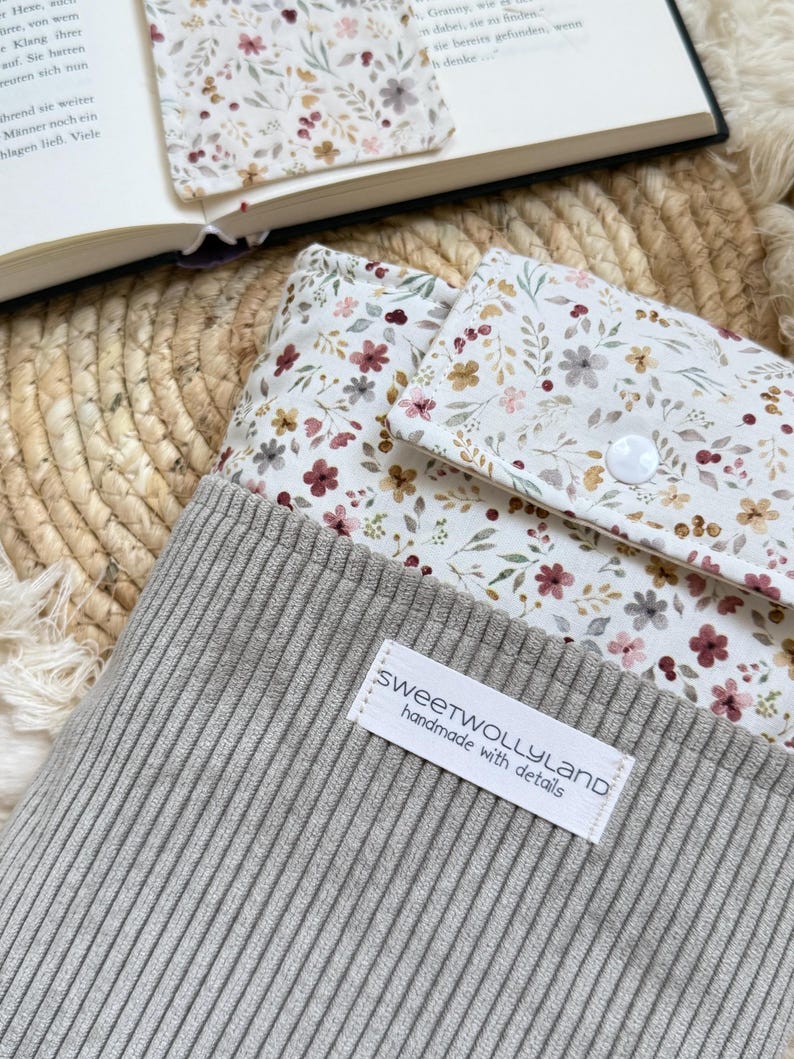 Ein grauer Beutel aus Rippenstoff mit einer blumig gemusterten Klappe und einem weißen Druckknopfverschluss. Auf der Vorderseite ist ein weißes rechteckiges Etikett mit dem schwarzen Text "sweetwollyland handmade with details" aufgenäht.