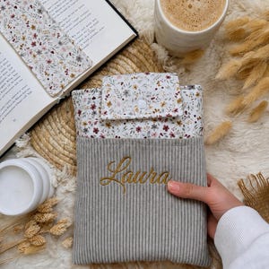 Personalisierte Buchhülle aus grauem Cord mit floralem Stoff und Namen Laura, handgemachter Buchschutz für Taschenbücher, Hardcover und Bücherliebhaber.