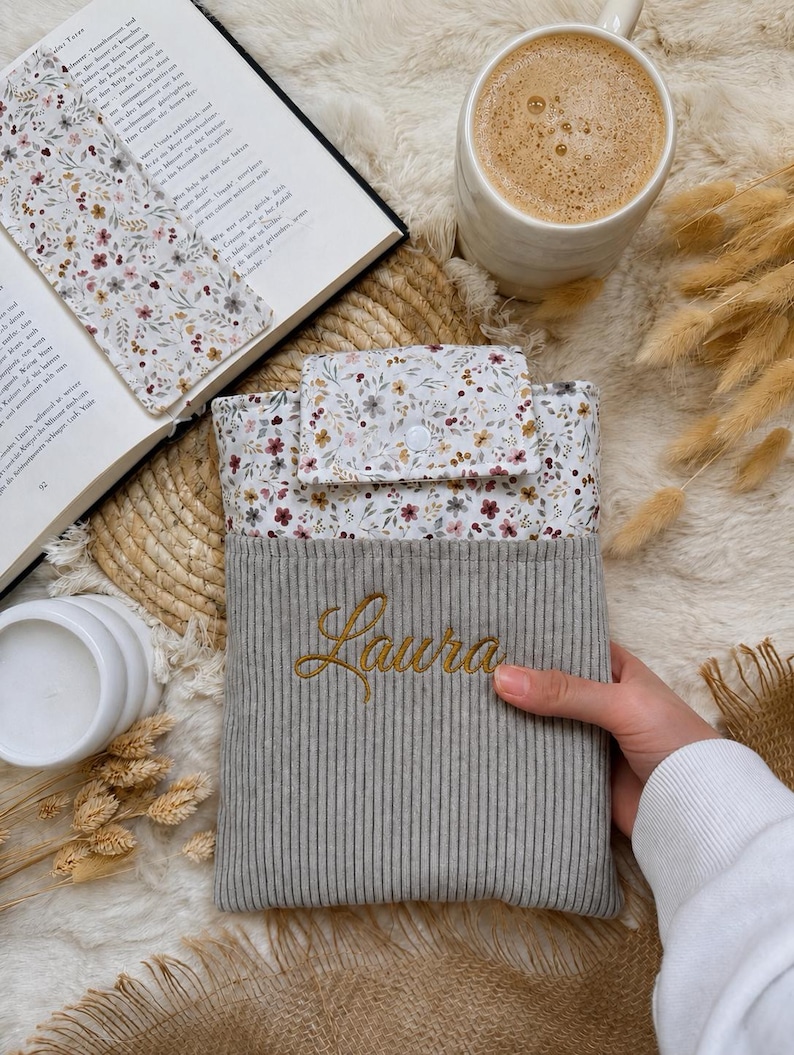 Personalisierte Buchhülle aus grauem Cord mit floralem Stoff und Namen Laura, handgemachter Buchschutz für Taschenbücher, Hardcover und Bücherliebhaber.