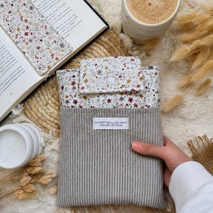 Eine rechteckige Buchhülle aus Stoff mit einem unteren Teil aus grauem Ripp-Cord und einer floral gemusterten Klappe mit weißem Druckknopf. Ein weißes Etikett auf der Vorderseite zeigt den Text sweetwollyland handmade with details.
