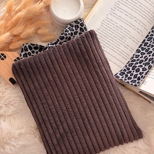 Eine braune Cord Buchhülle  steht im Mittelpunkt, mit einem Leopardenmuster-Band und innen Tasche , das teilweise zu sehen ist. Ein weißer Keramik-Kerzenhalter und ein Buch im Hintergrund schaffen eine gemütliche Atmosphäre.