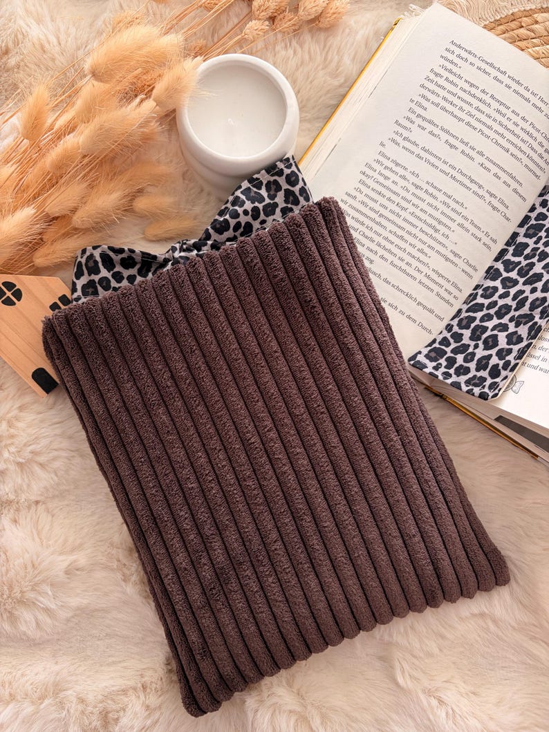 Eine braune Cord Buchhülle  steht im Mittelpunkt, mit einem Leopardenmuster-Band und innen Tasche , das teilweise zu sehen ist. Ein weißer Keramik-Kerzenhalter und ein Buch im Hintergrund schaffen eine gemütliche Atmosphäre.