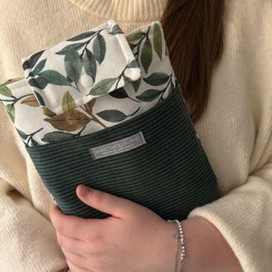 Könnte beinhalten: Eine rechteckige Stofftasche mit einem unteren Teil aus dunkelgrünem Kord und einer weißen oberen Klappe mit grün-braunem Blattmuster. Ein weißer Druckknopf verschließt die Klappe. Ein graues Etikett trägt die Aufschrift SWEETWOLLYLAND.