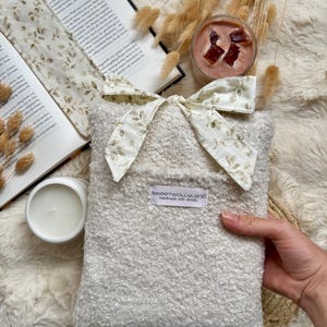 Könnte beinhalten: Eine rechteckige, cremefarbene Tasche aus strukturiertem Sherpa-Stoff mit einer weißen Stoffschleife mit zartem braunem Blattmuster an der Oberseite. Ein kleines weißes Etikett auf der Vorderseite zeigt den Text "sweetwollyland handmade with details".
