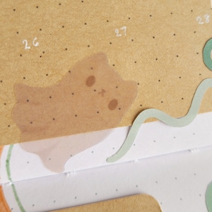 Może przedstawiać: Zbliżenie strony planera z naklejką z jasnobrązowym kotem. Naklejka ma przezroczystą nakładkę i jest umieszczona na kropkowanym, brązowym tle. Widoczne są białe cyfry 26, 27 i 28. Obecny jest również zielony, falisty element papieru.